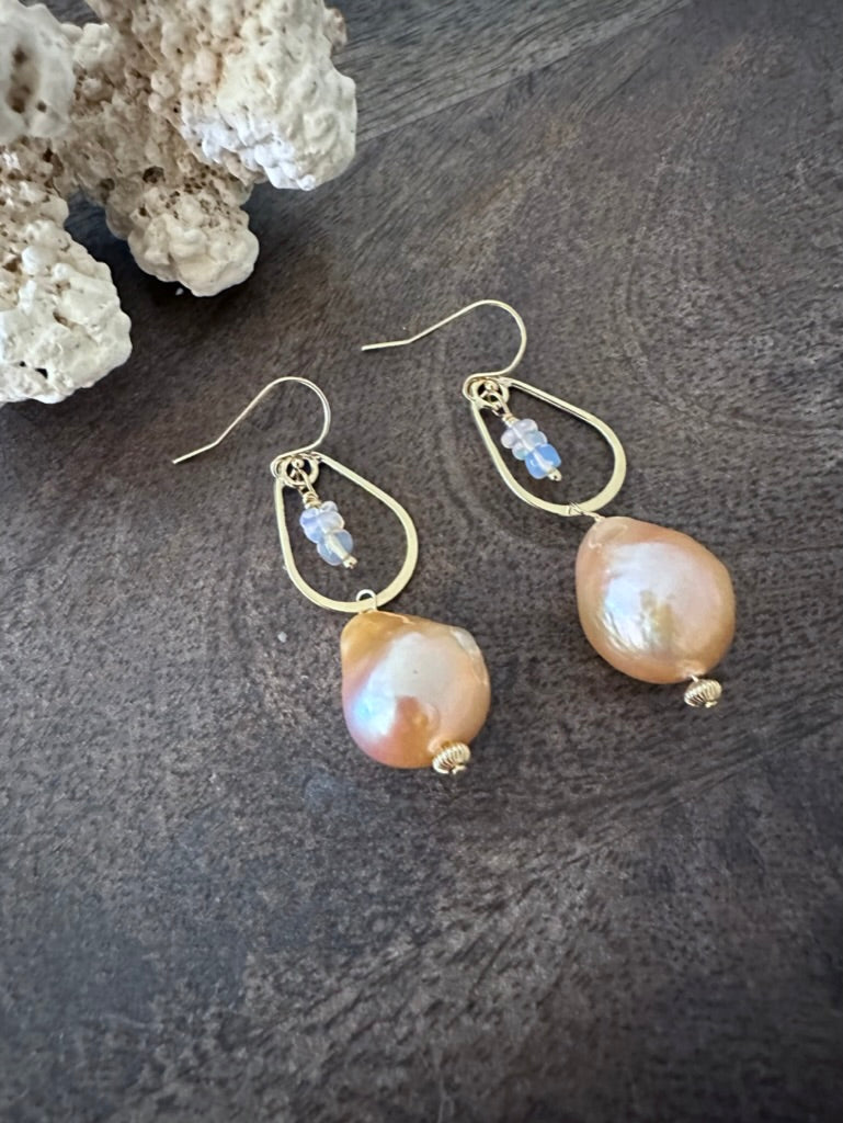 a pair of earrings on a wooden background with white coral in the upper left corner. There are little opal beads hanging inside of a golden teardrop. off of that is a large peachy fireball shaped pearl