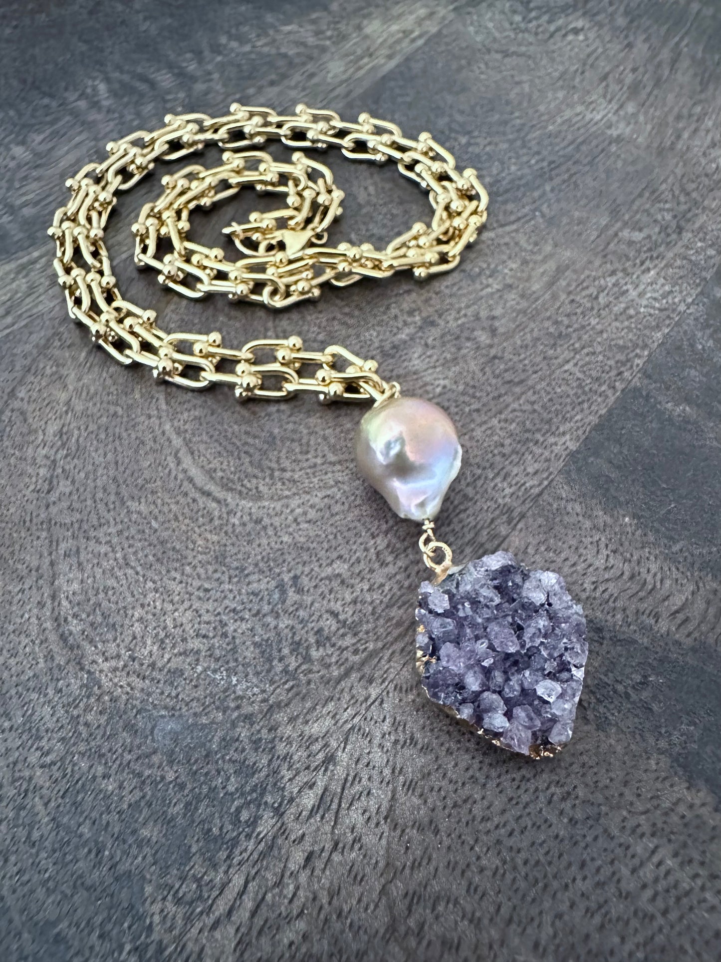 Large pink fireball shaped pearl woth a purple druzy pendant on a coiled gold chain on a wooden background