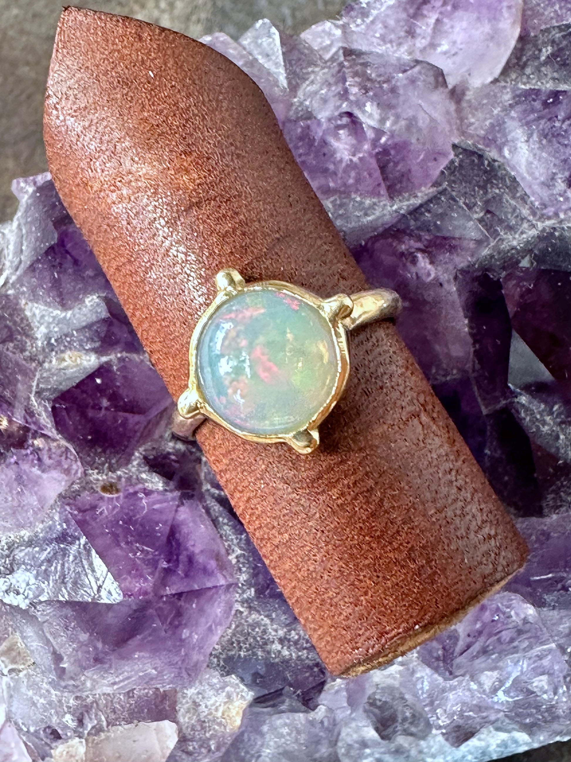 a round opal ring in gold iwth four beads at the four directins is on a piece of brown leather sitting on a piece of amethyst on grey wood