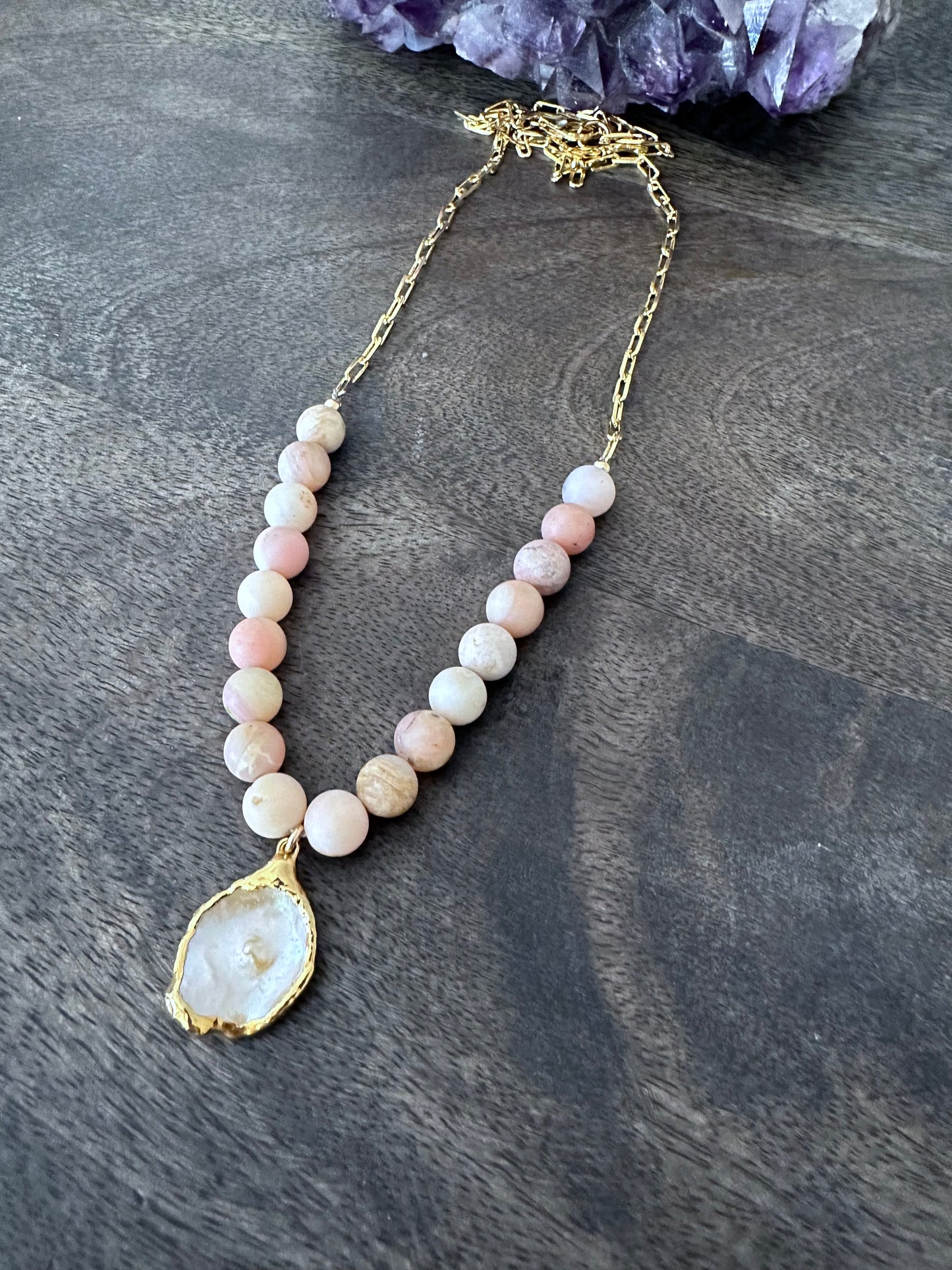 a necklace on a grey wooden background with pink beads and gold chain. theres is a white flat pearl as the center pendant