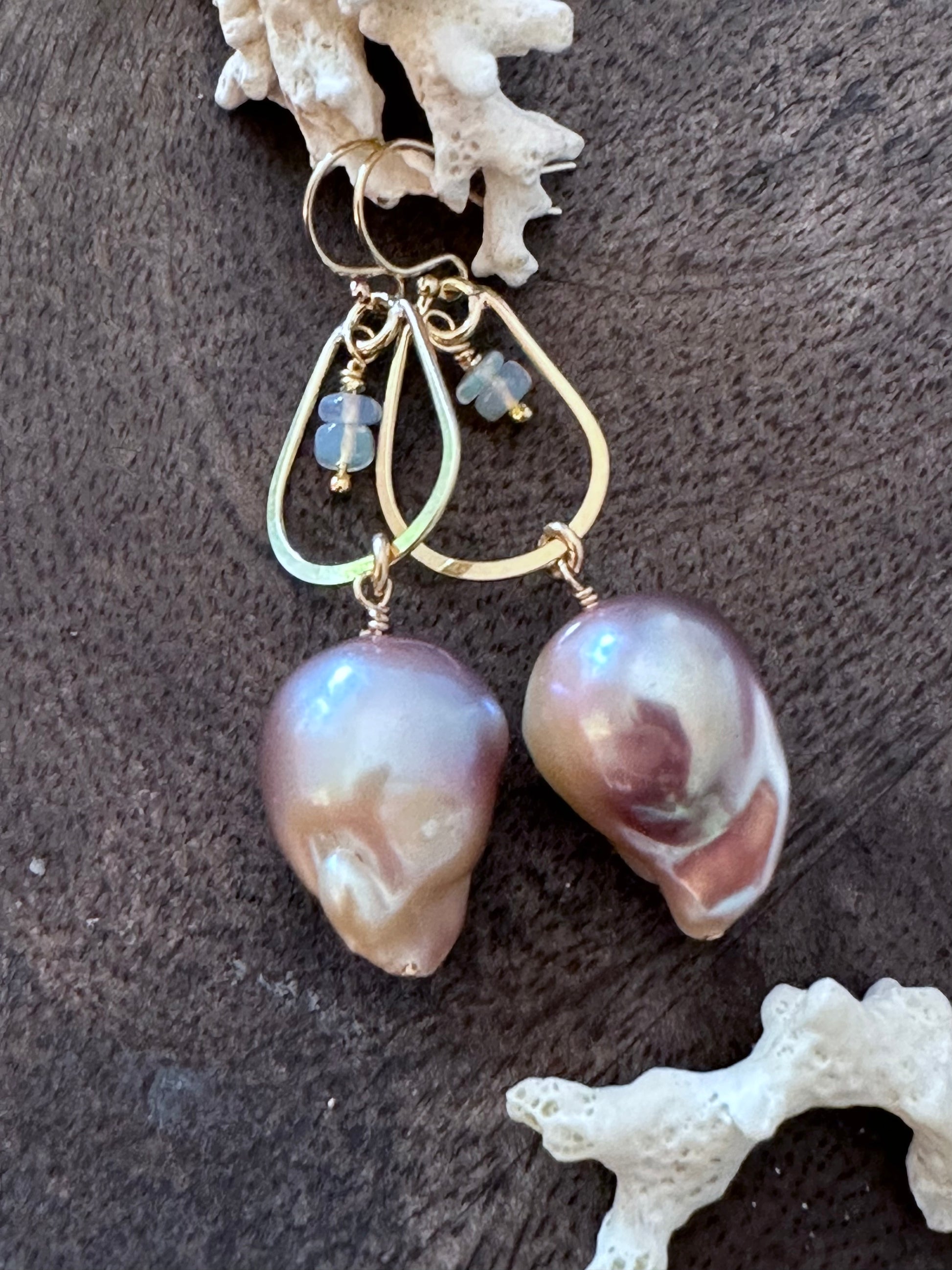 a pair of earrings on a wooden background with white coral in the upper left corner. There are little opal beads hanging inside of a golden teardrop. off of that is a large purple fireball shaped pearl