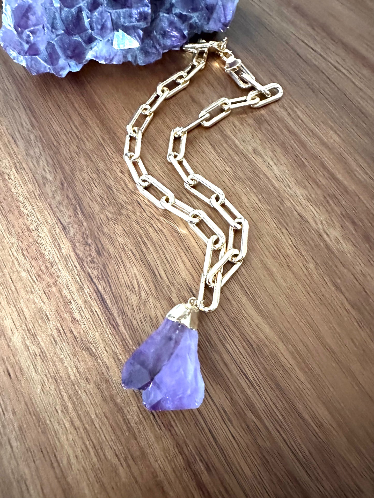 a large double pointed amethyst purple crystal pendant on a large gold chain on a wooden background. a large amethyst crystal sits at the top of the image