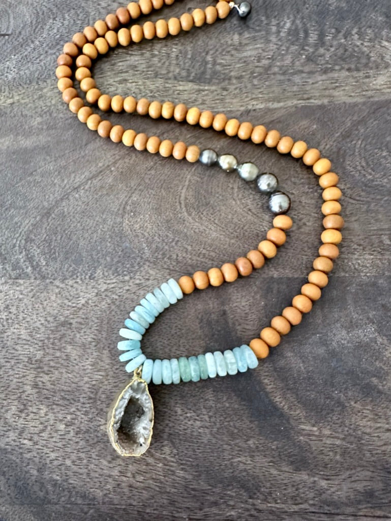a necklace with blue aquamarine, sandalwood and tahtian pearl beads with a raw geode is on a wooden background
