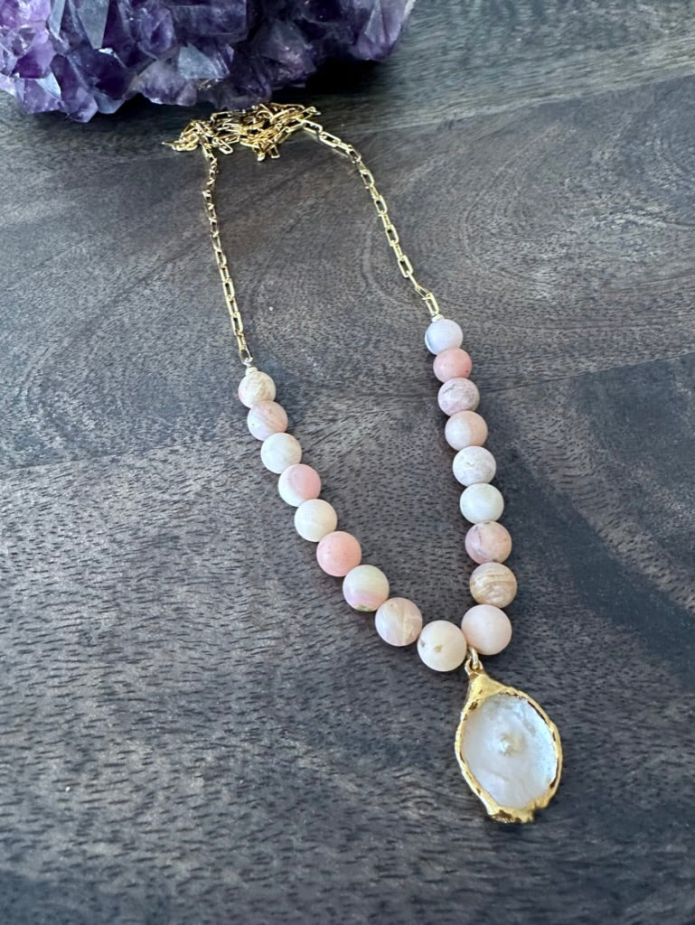 a necklace on a grey wooden background with pink beads and gold chain. theres is a white flat pearl as the center pendant