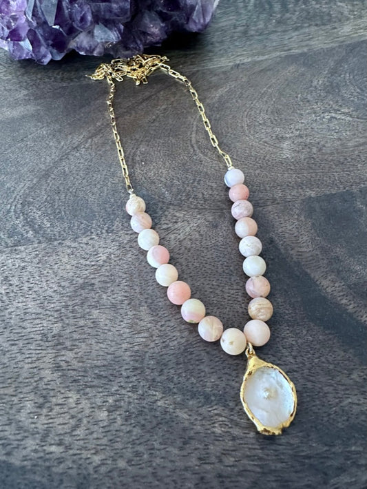  a necklace on a grey wooden background with pink beads and gold chain. theres is a white flat pearl as the center pendant