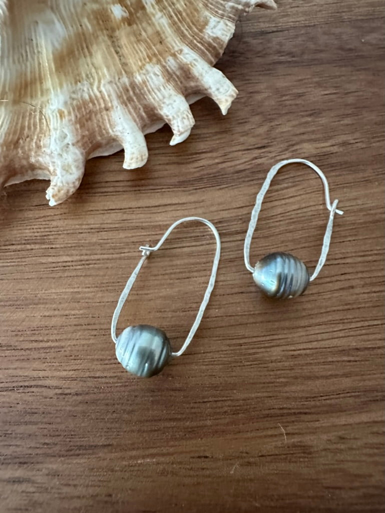 a pair of earrings on a wooden board with a shell in the upper left corner. they are ovoal shaped silvder wire with black pearls with ridges on them