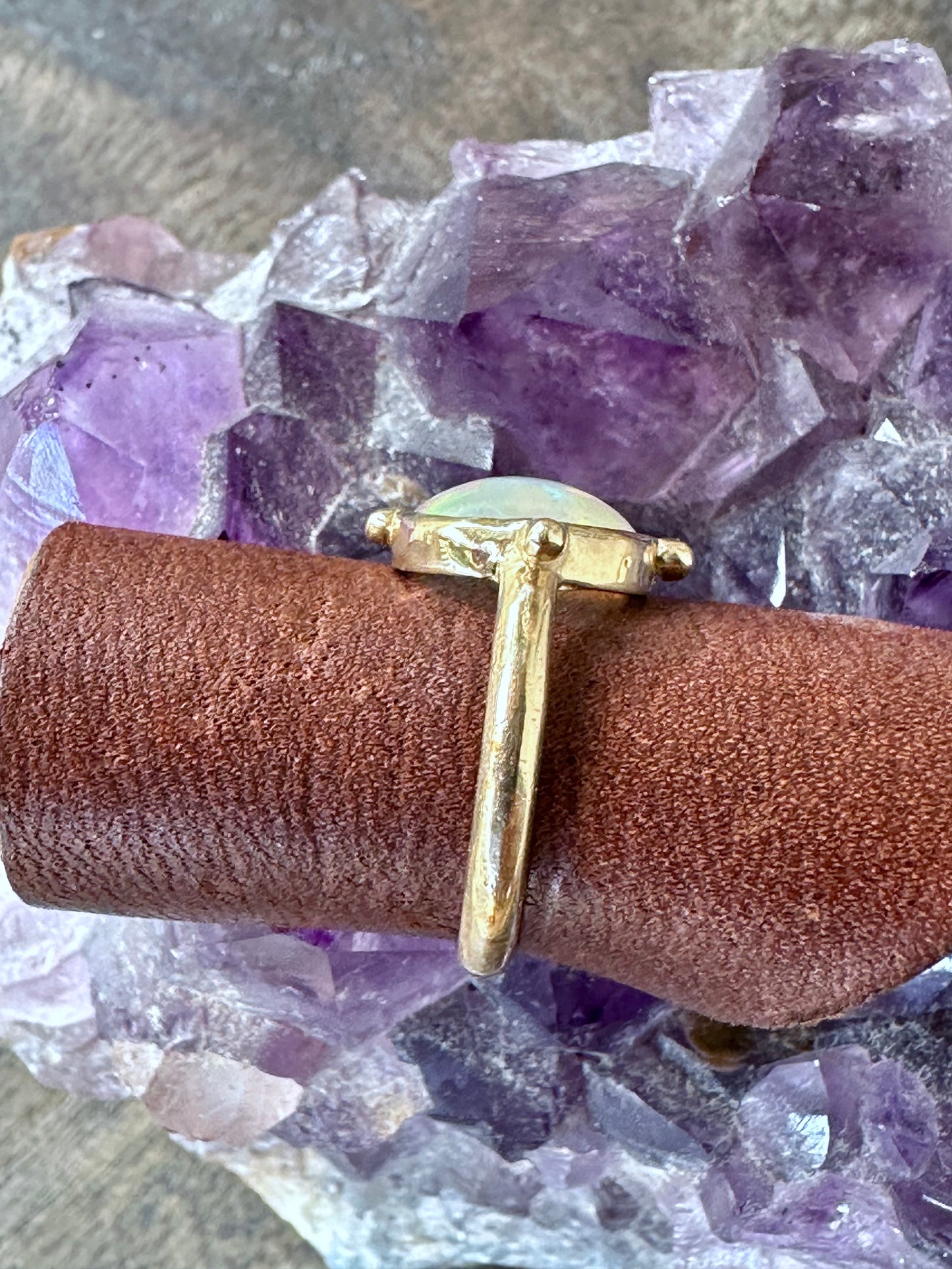 a side view of a round opal ring in gold iwth four beads at the four directins is on a piece of brown leather sitting on a piece of amethyst on grey wood