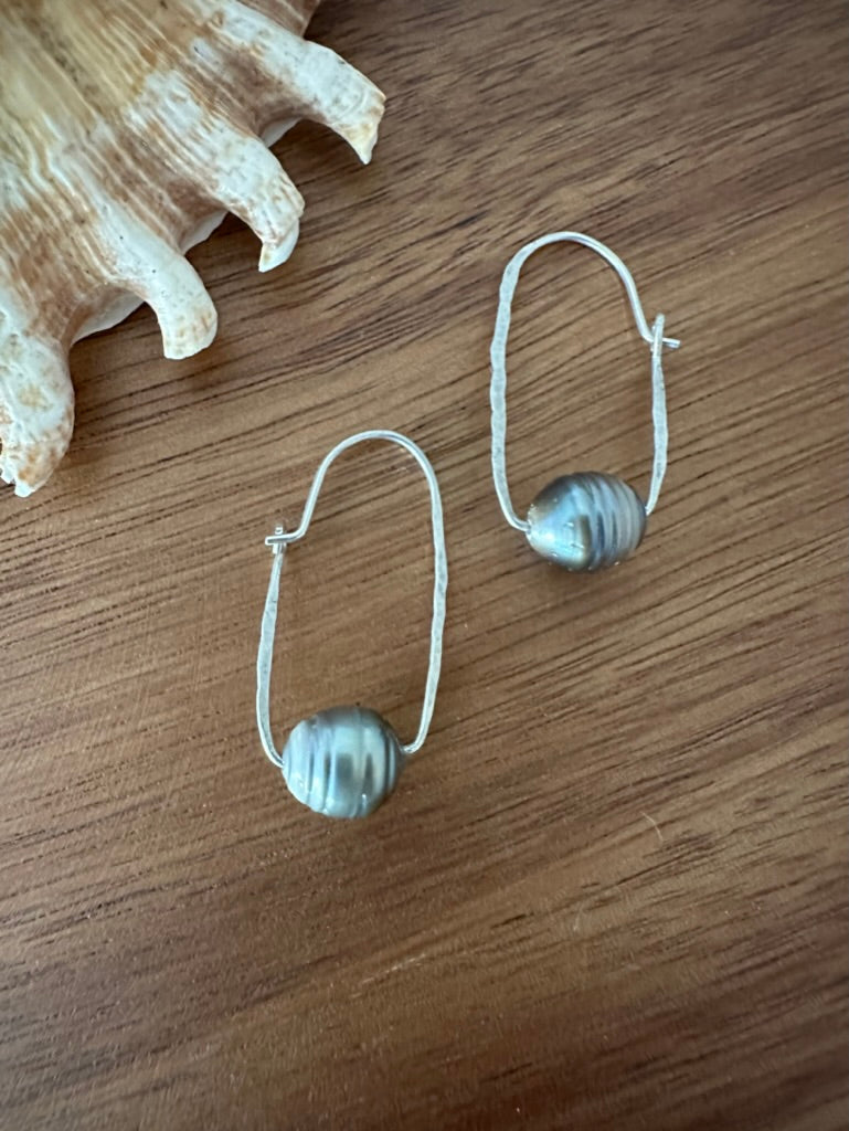 a pair of earrings on a wooden board with a shell in the upper left corner. they are ovoal shaped silvder wire with black pearls with ridges on them