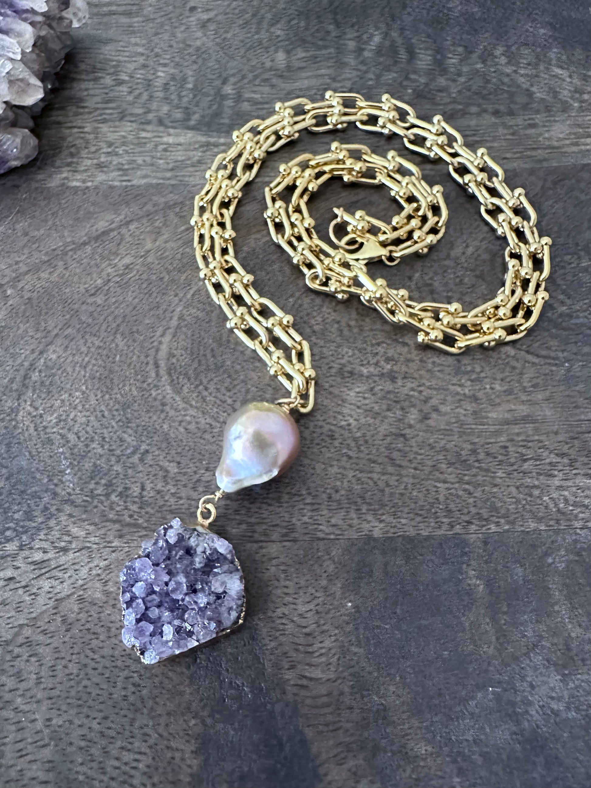 Large pink fireball shaped pearl woth a purple druzy pendant on a coiled gold chain on a wooden background