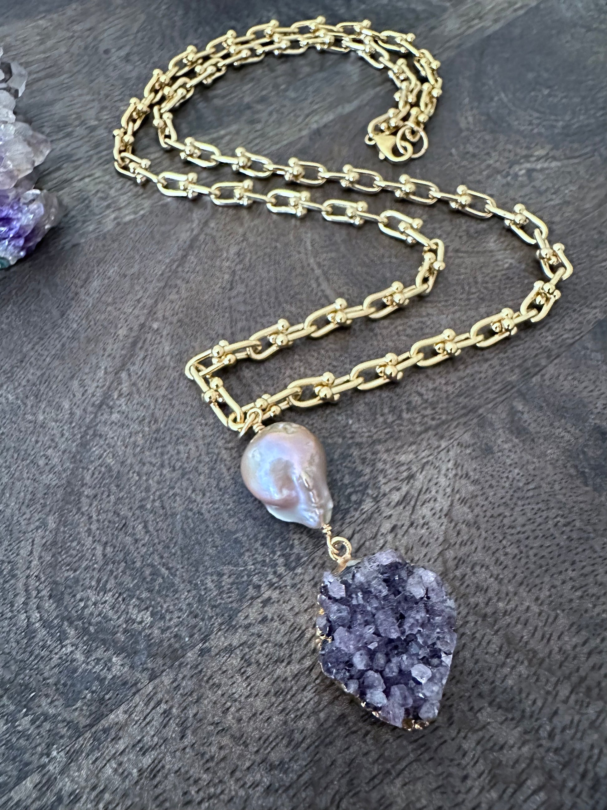 Large pink fireball shaped pearl woth a purple druzy pendant on a coiled gold chain on a wooden background