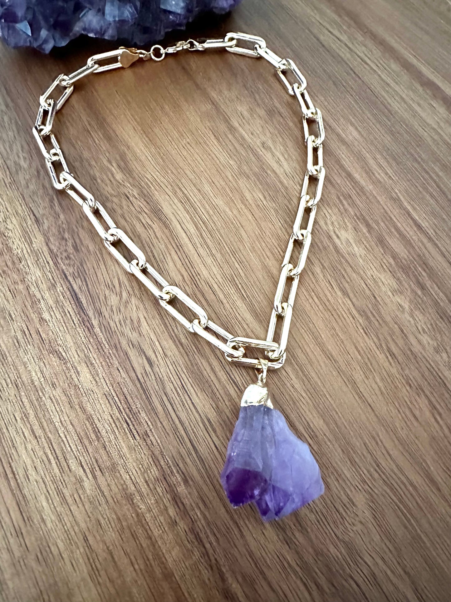  a large double pointed amethyst purple crystal pendant on a large gold chain on a wooden background. a large amethyst crystal sits at the top of the image