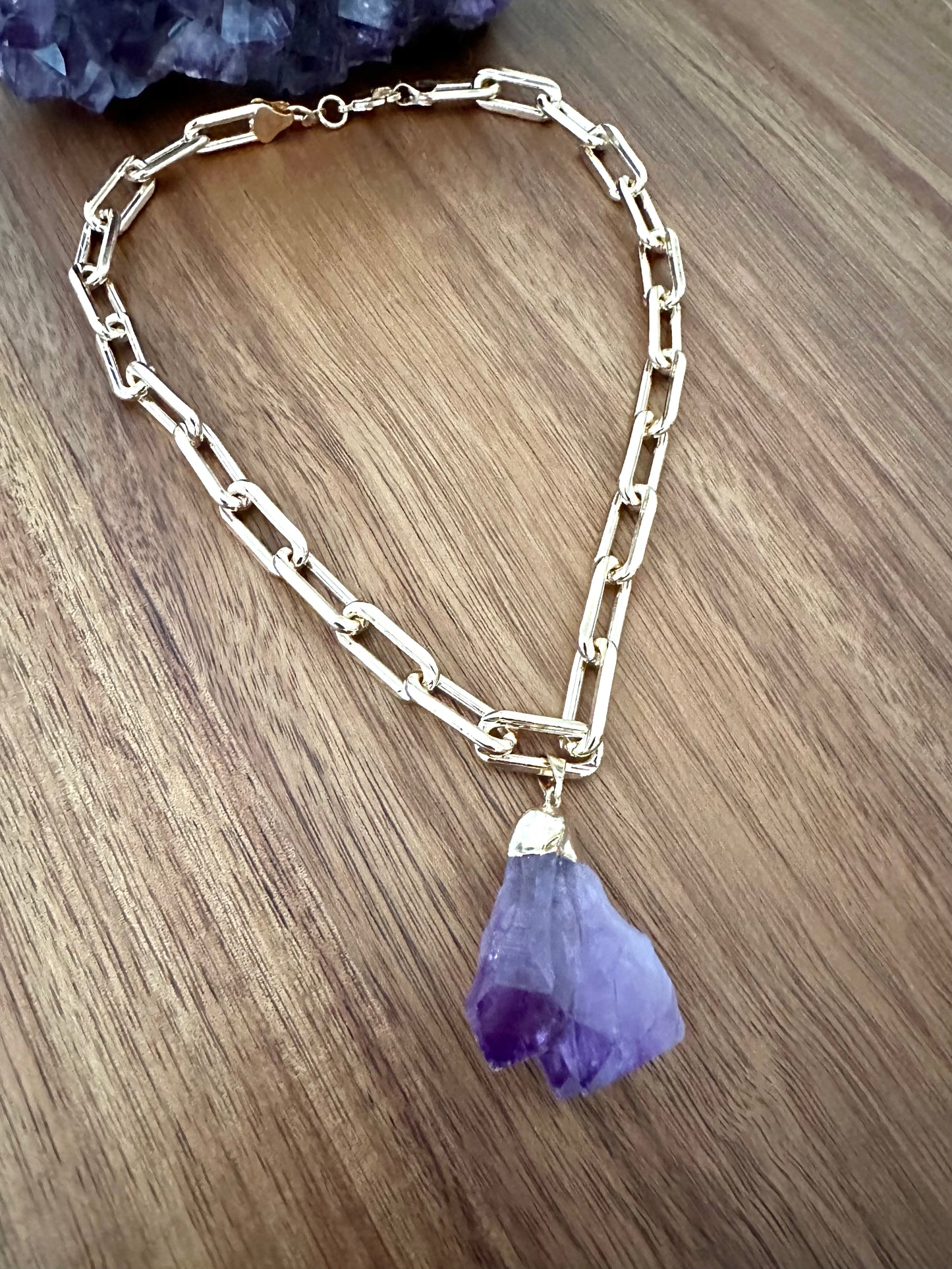 a large double pointed amethyst purple crystal pendant on a large gold chain on a wooden background. a large amethyst crystal sits at the top of the image