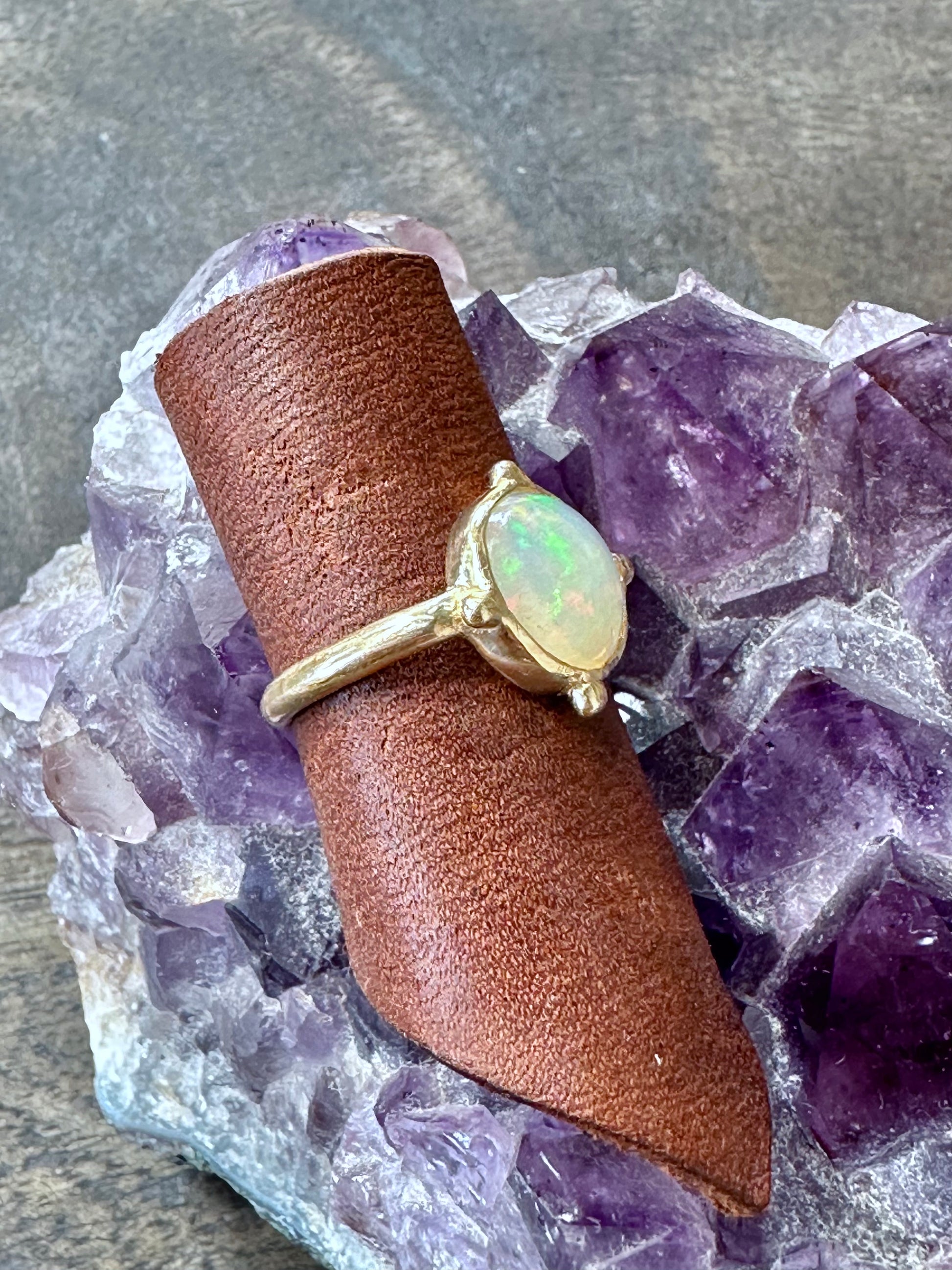 a 3/4 view of a round opal ring in gold iwth four beads at the four directins is on a piece of brown leather sitting on a piece of amethyst on grey wood