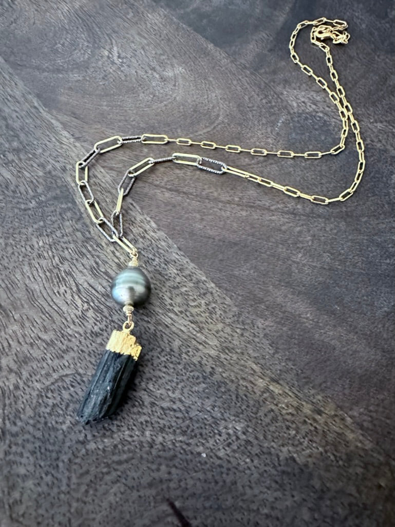 a black raw tourmaline and tahitian pearl pendand on a mixed gold and silver paperclip chain on a wooden background