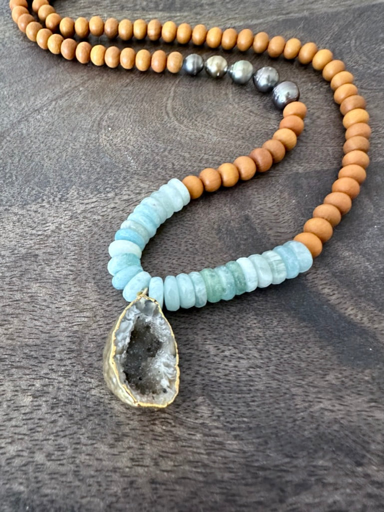 a necklace with blue aquamarine, sandalwood and tahtian pearl beads with a raw geode is on a wooden background