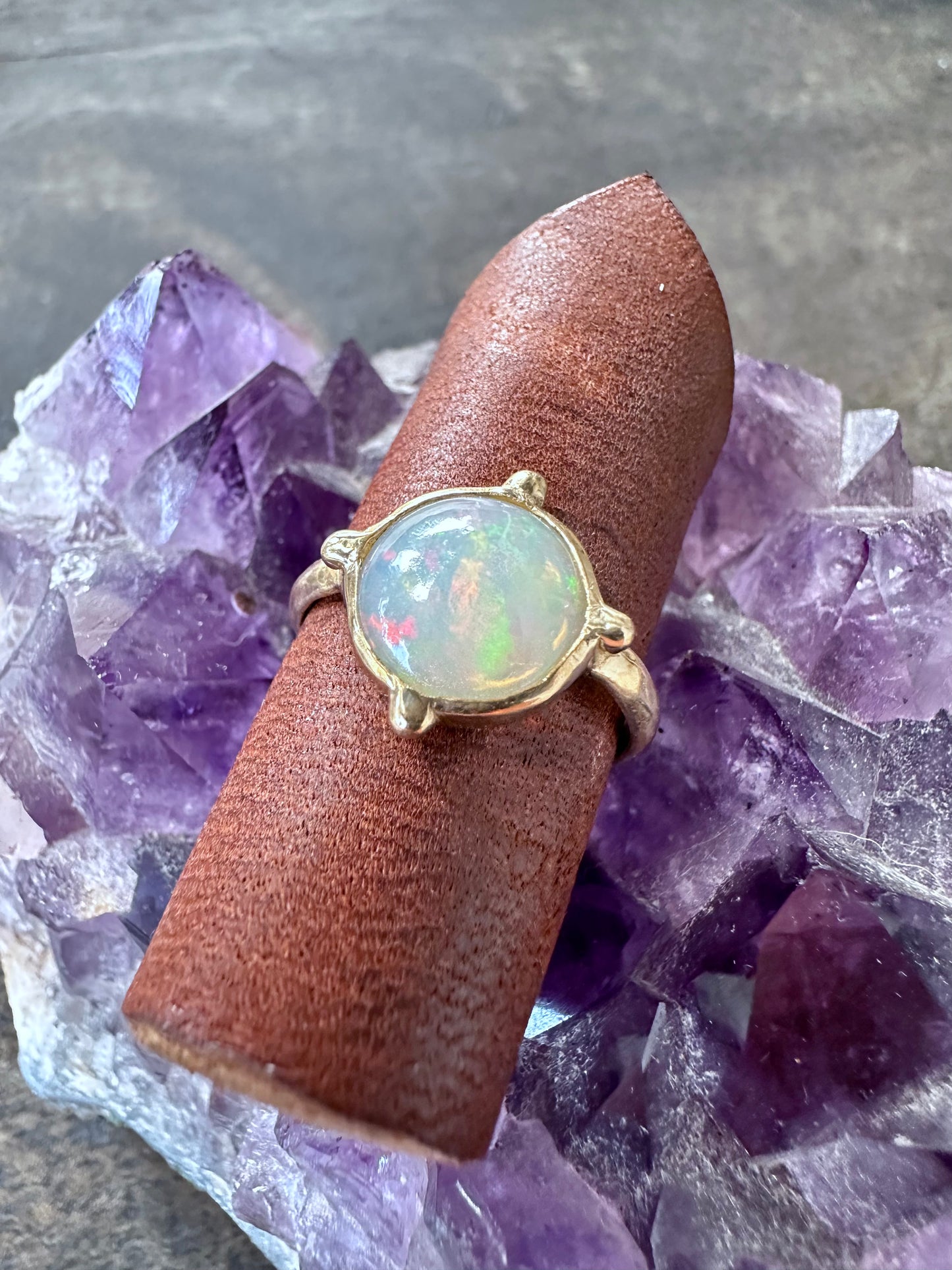 a round opal ring in gold iwth four beads at the four directins is on a piece of brown leather sitting on a piece of amethyst on grey wood