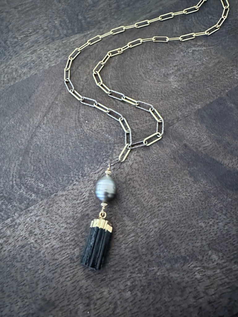 a black raw tourmaline and tahitian pearl pendand on a mixed gold and silver paperclip chain on a wooden background