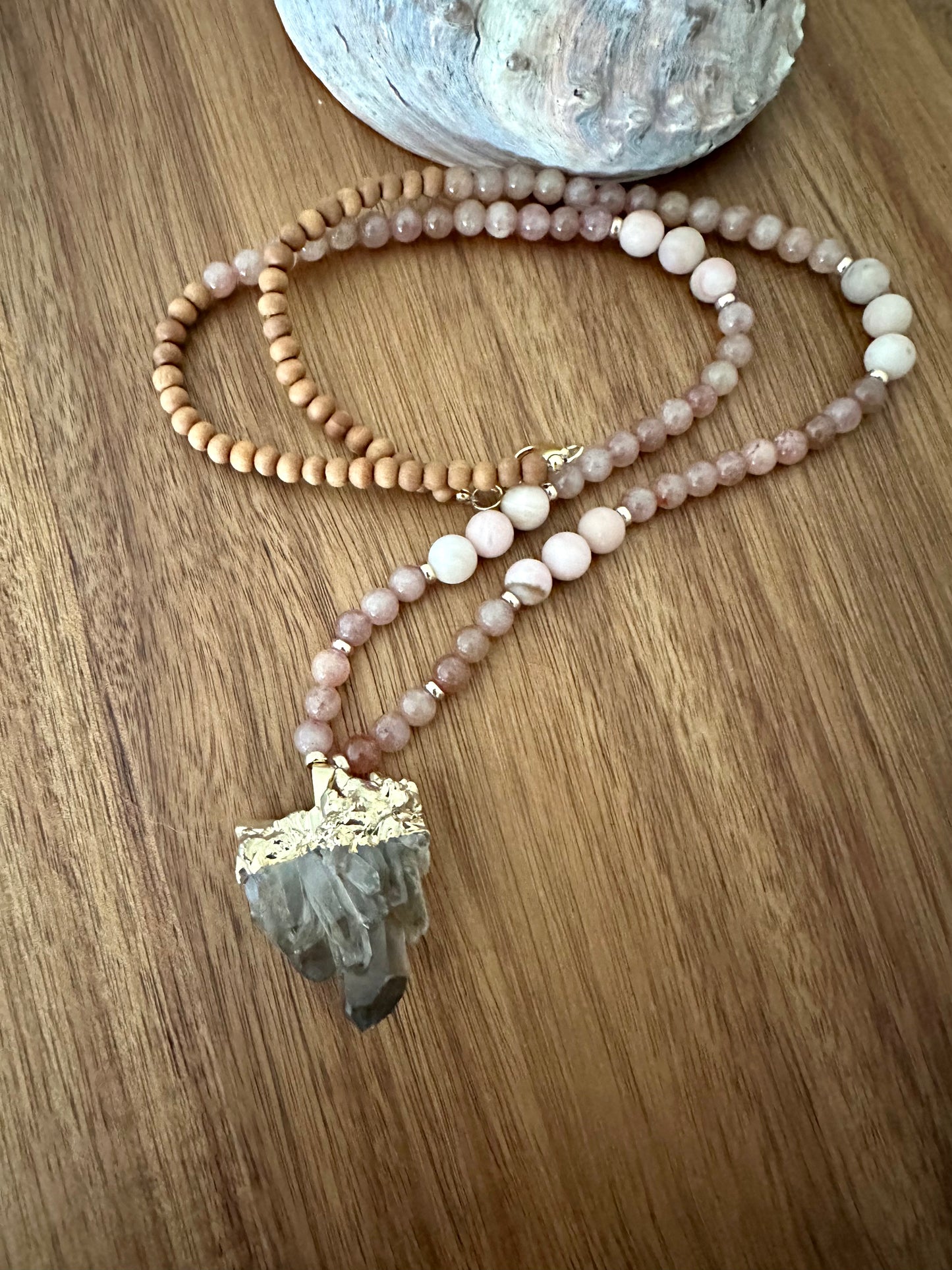 a necklace semi coiled up with pink stone beads, and wooden beads with a large smokey quartz pendant on a wooden board with an abalone shell in the background