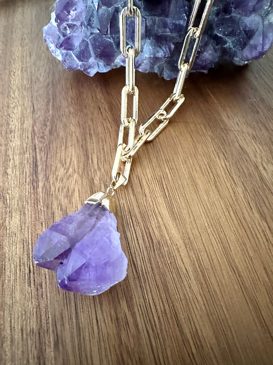 a large double pointed amethyst purple crystal pendant on a large gold chain on a wooden background. a large amethyst crystal sits at the top of the image
