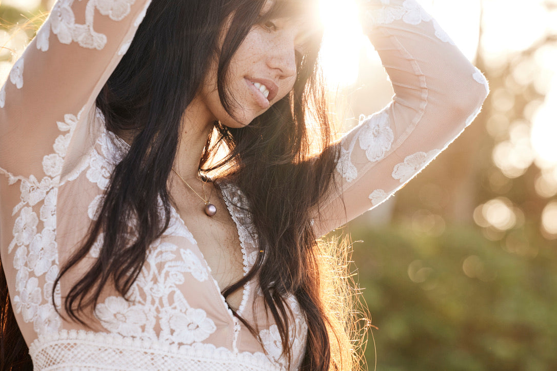 a beautiful brown haired woman in a lacy dress in front of sunrays. She is smailing and wearing a pinkish pearl necklace
