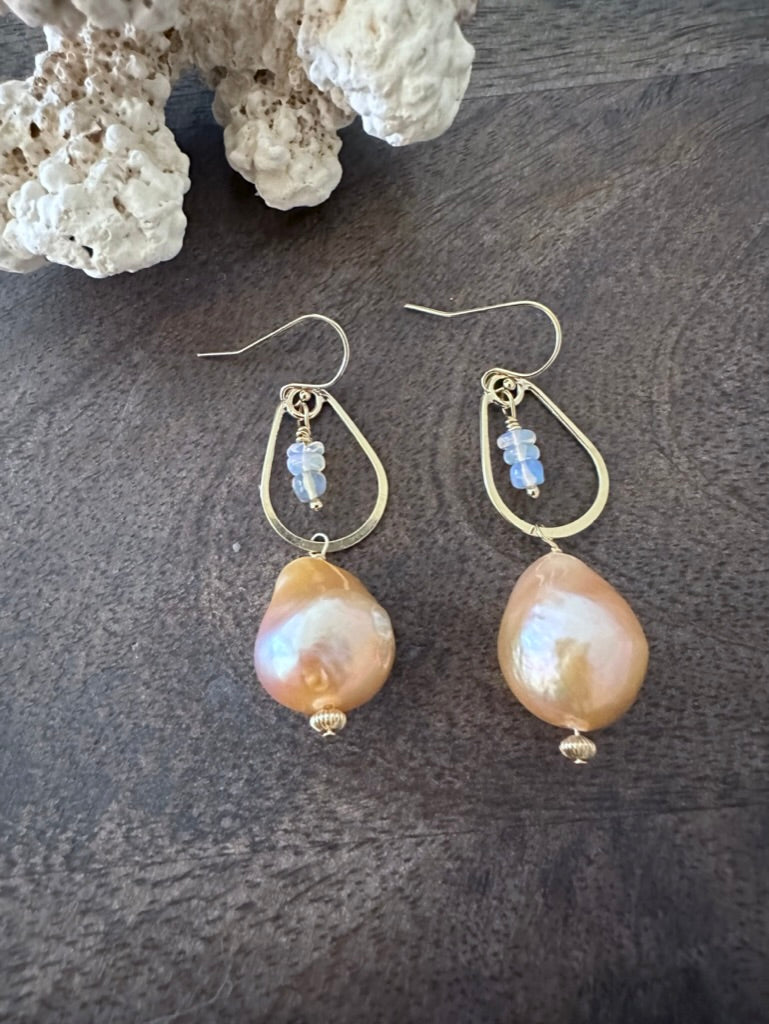 a pair of earrings on a wooden background with white coral in the upper left corner. There are little opal beads hanging inside of a golden teardrop. off of that is a large peachy fireball shaped pearl