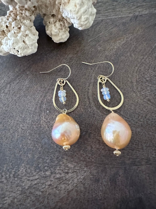 a pair of earrings on a wooden background with white coral in the upper left corner. There are little opal beads hanging inside of a golden teardrop. off of that is a large peachy fireball shaped pearl 