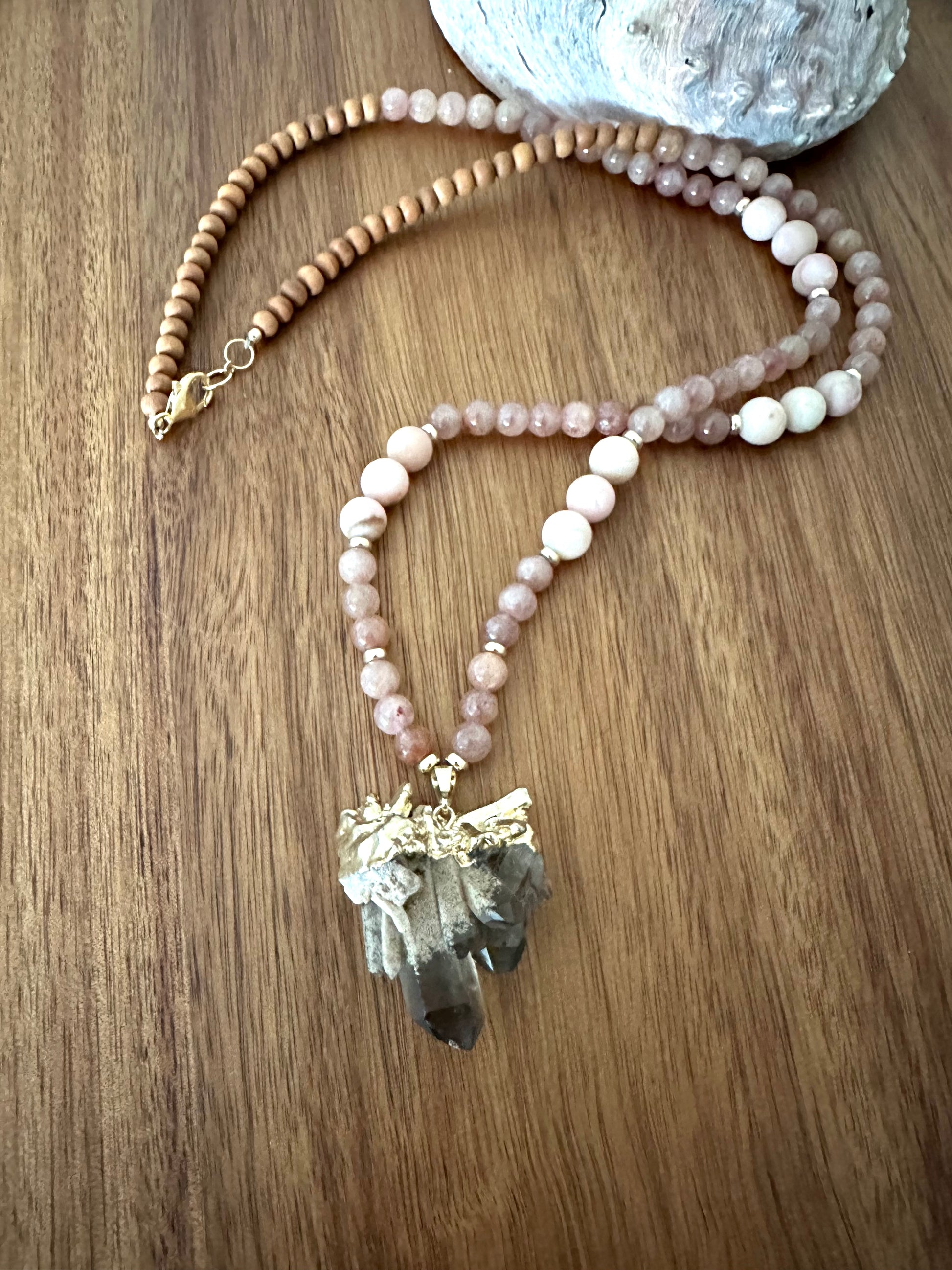 a necklace semi coiled up with pink stone beads, and wooden beads with a large smokey quartz pendant on a wooden board with an abalone shell in the background