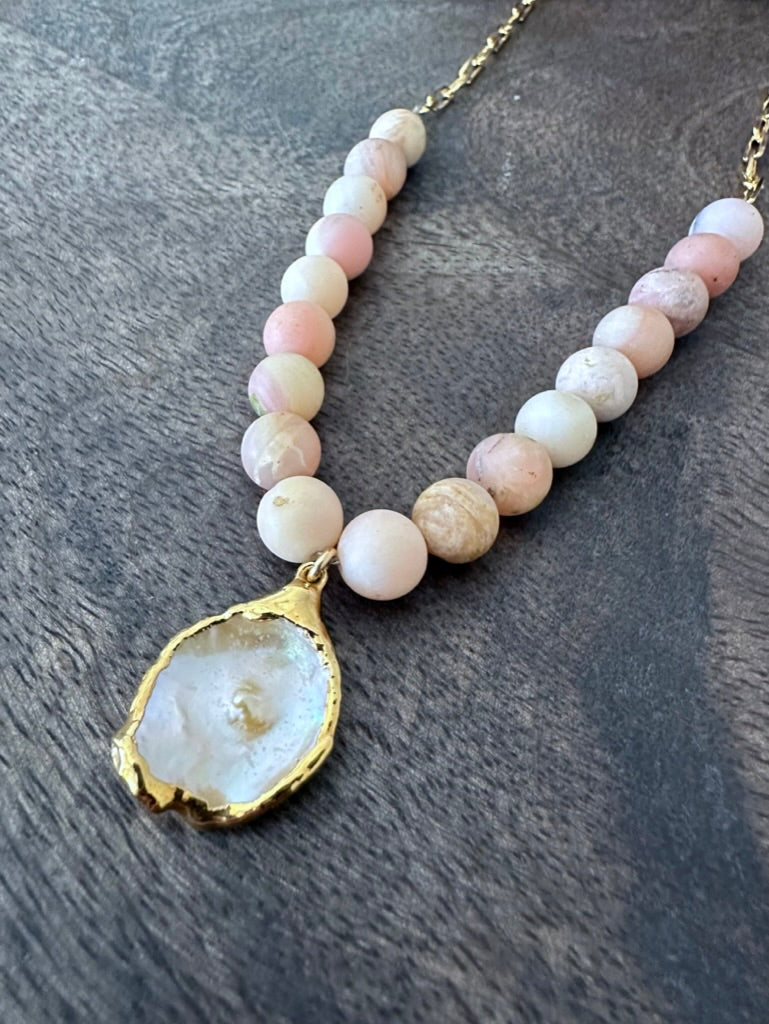 close up of a necklace on a grey wooden background with pink beads and gold chain. theres is a white flat pearl as the center pendant