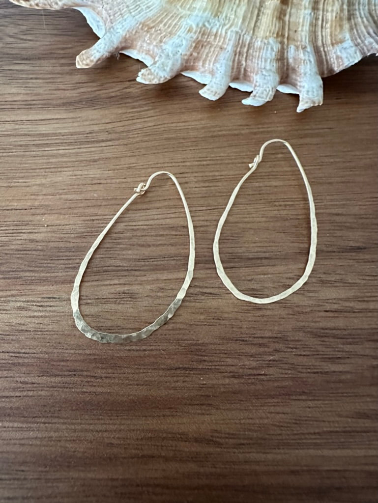 a pair of gold hoop earrings that are hammered and made from a single continuous wire. they are shaped like a lare teardrop or kind of like and avocado. they sit on a wooden backdrop with a shell in the upper part of the frame