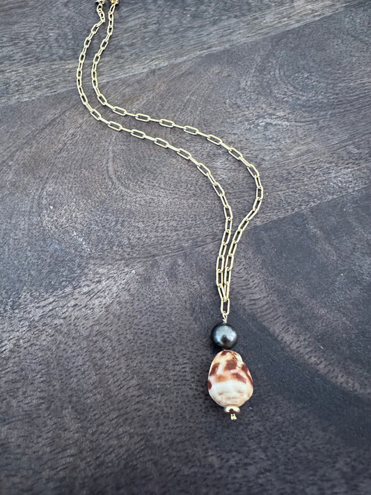 a brown and shite spotted cone shell with a tahitian pearl aon a gold paperclip chain on a wooden background