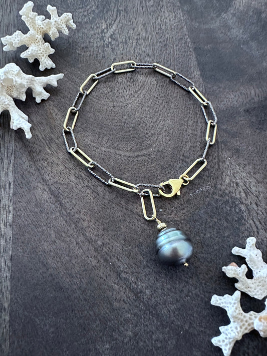 mixed silver and gold bracelet with a large teardrop tahtian black pearl with ridges lays in a circle on a woodedn background with white coralin the upper left and lower right corners