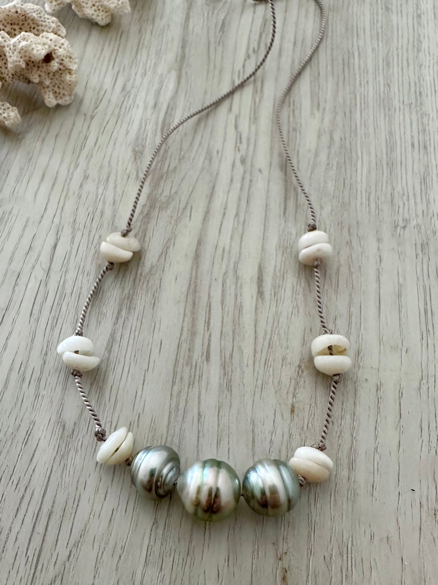 a necklace with three silvery tahitain pearls on a gry silk thread with white small puka shells is on a light grey wooden background. there is a small chink of coral in the upper left corner