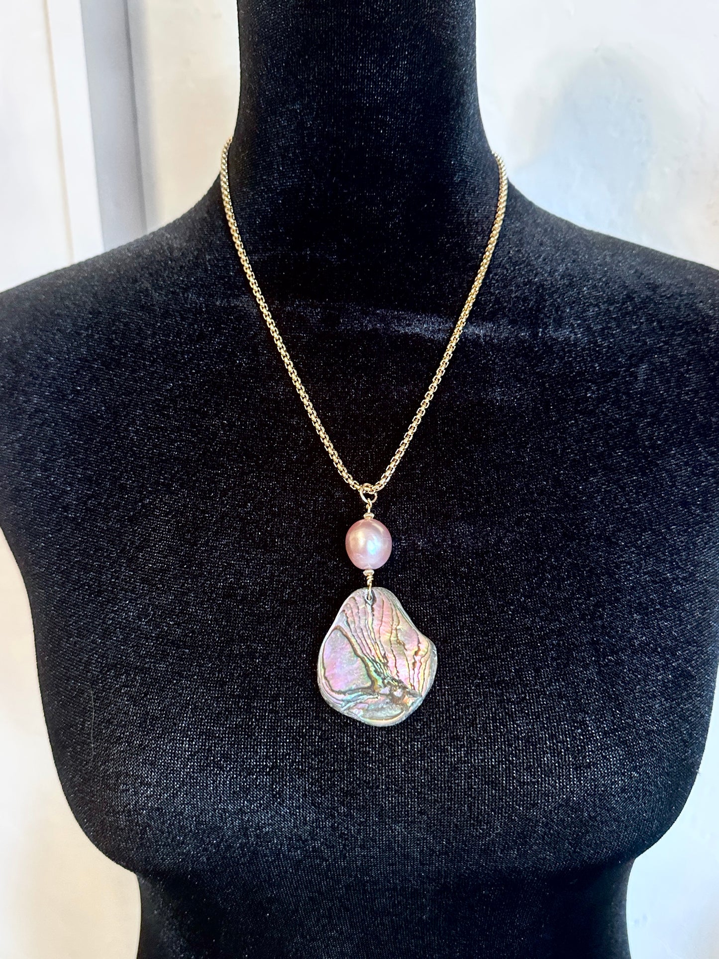 A gold chain necklace with a large, irregular iridescent abalone shell pendant and a small pink pearl bead, displayed on a black velvet mannequin bust.