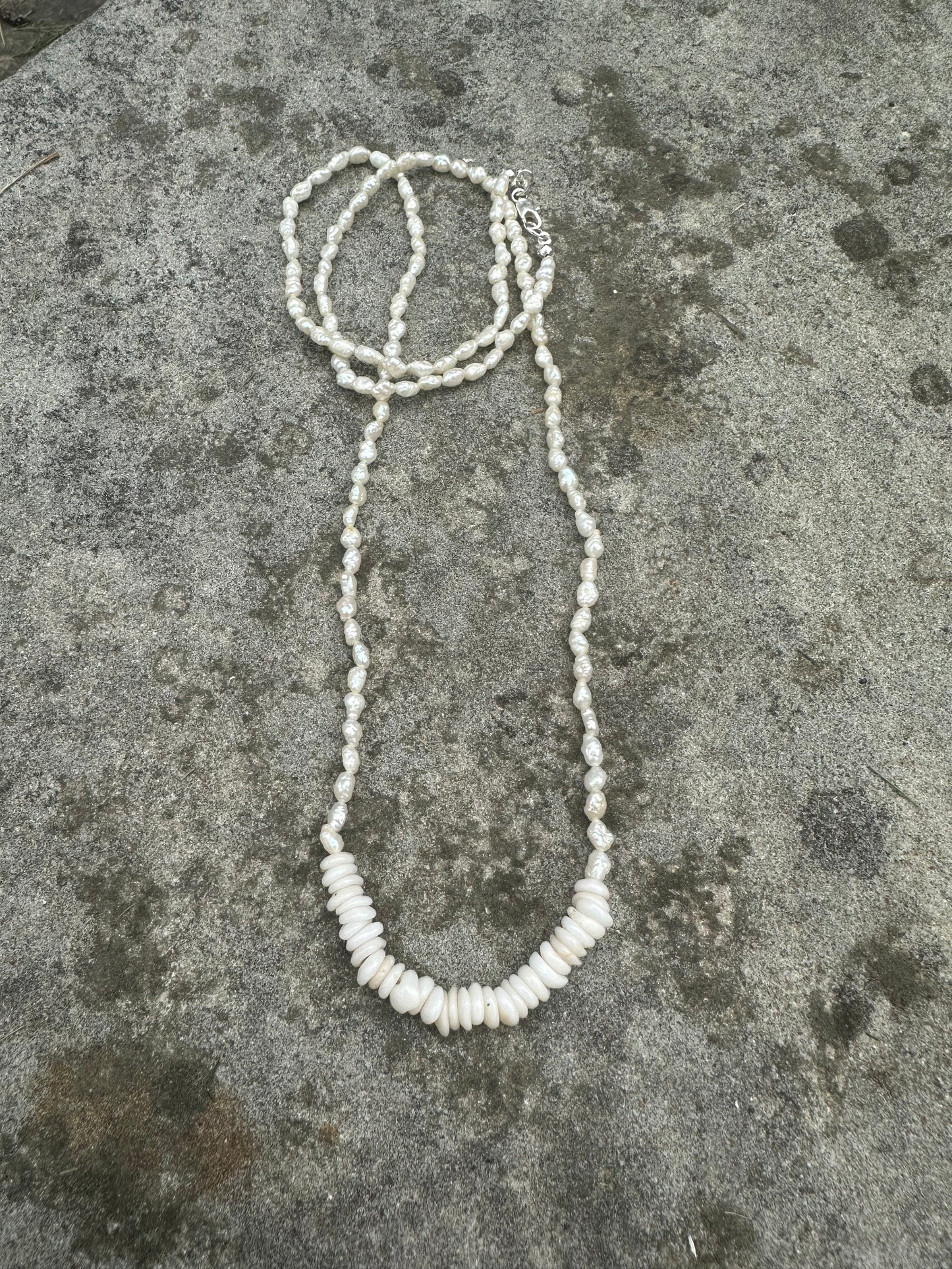 a long neckalce with white little pearls and a bar of white puka shells on a grey stone background.
