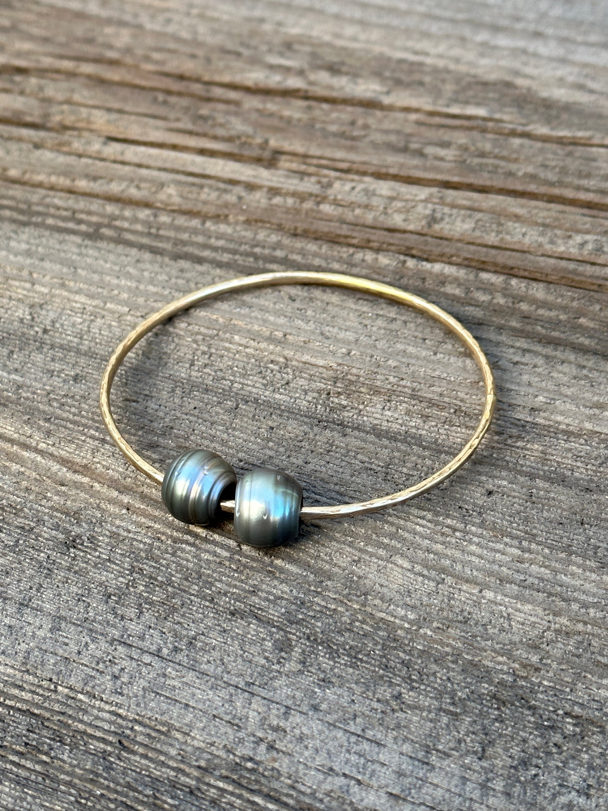 Two black baroque Tahitian pearls on a gold bangle resting on a wooden background