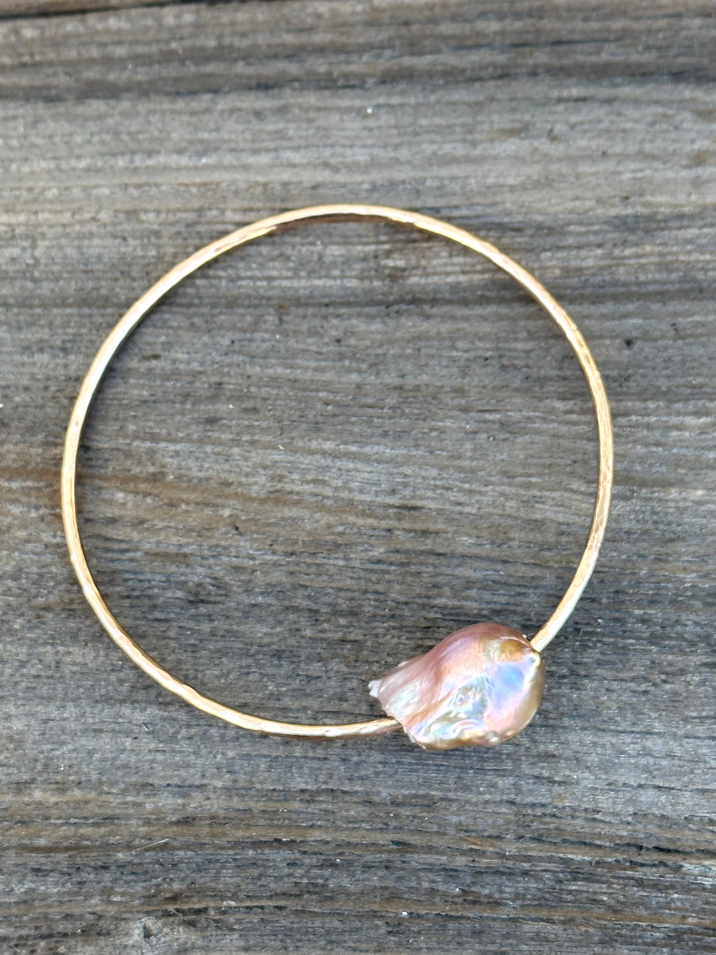 A gold bangle with a large oddly shaped fireball pearl in light hues of pink on a wooden background.