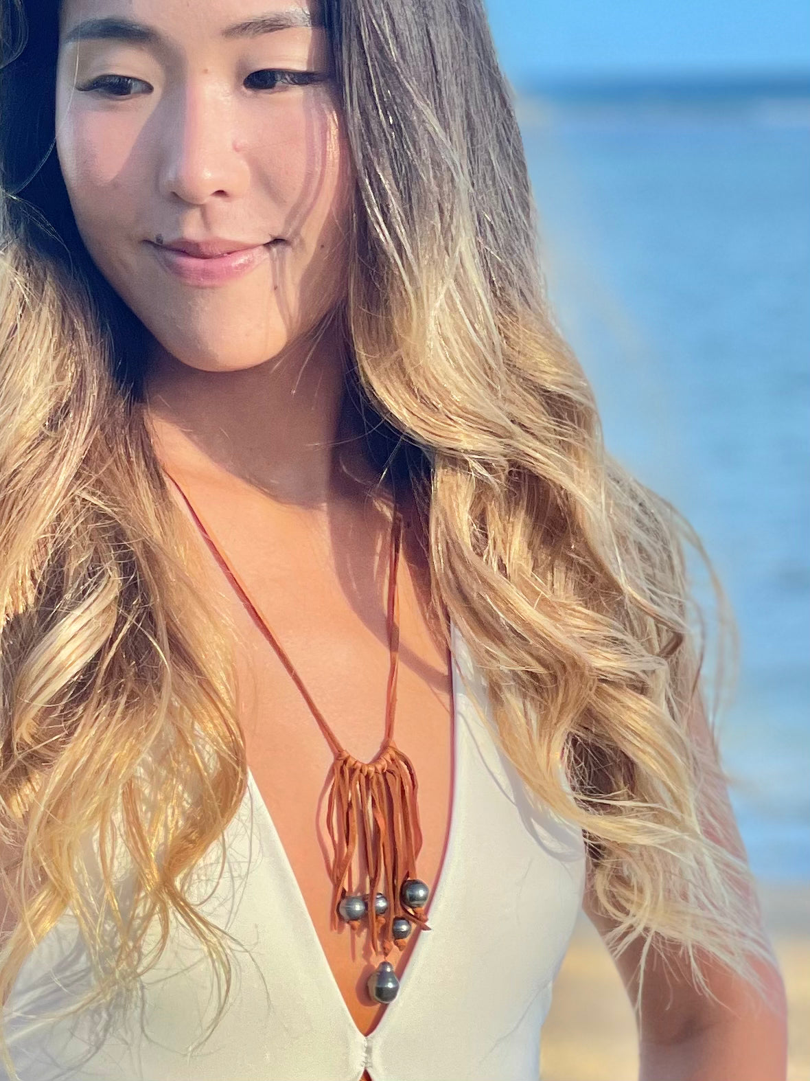 A pretty woman on a beach wearing a white bathing suit with a leather fringed necklace with dark black tahitian pearls