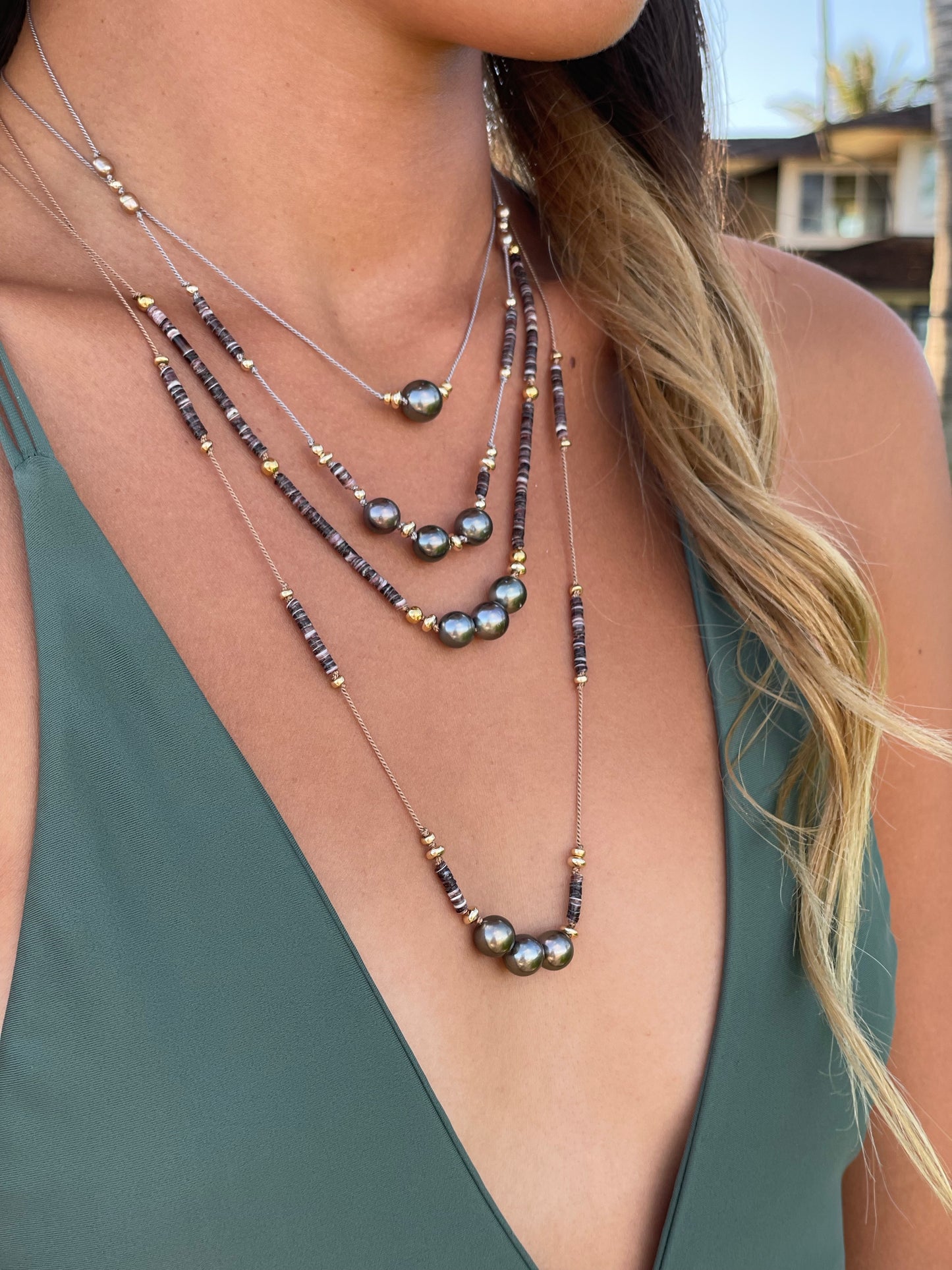 a woman in a green bikini, all you see is her chest and four necklaces layered. they have black pearls and brwonish beads with gold beads. her hair is golden colored and lays on her left shoulder. The first necklace has one pearl, the next three have three pearls.