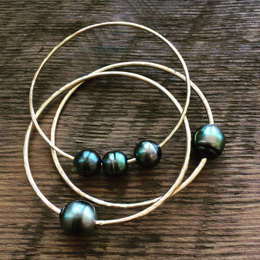 a stack of bangles with black pearls on them lays on a wooden background. two of them have a single bangle and the thied one has three pearls