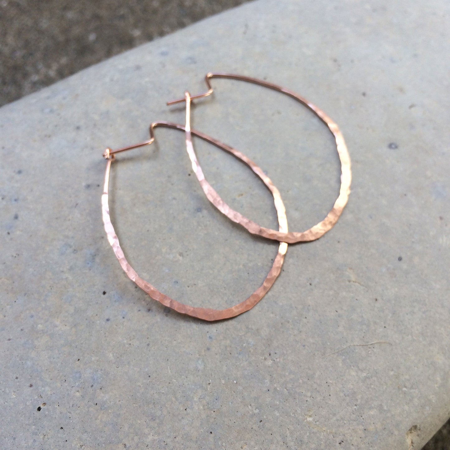 Two rose gold organically shaped hammered textured hoop earrings on a stone background