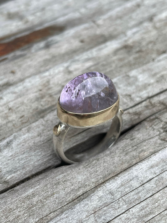 a large purple domes ring with a gold band around it and gold round accent on a silver band sits in some grey wood.