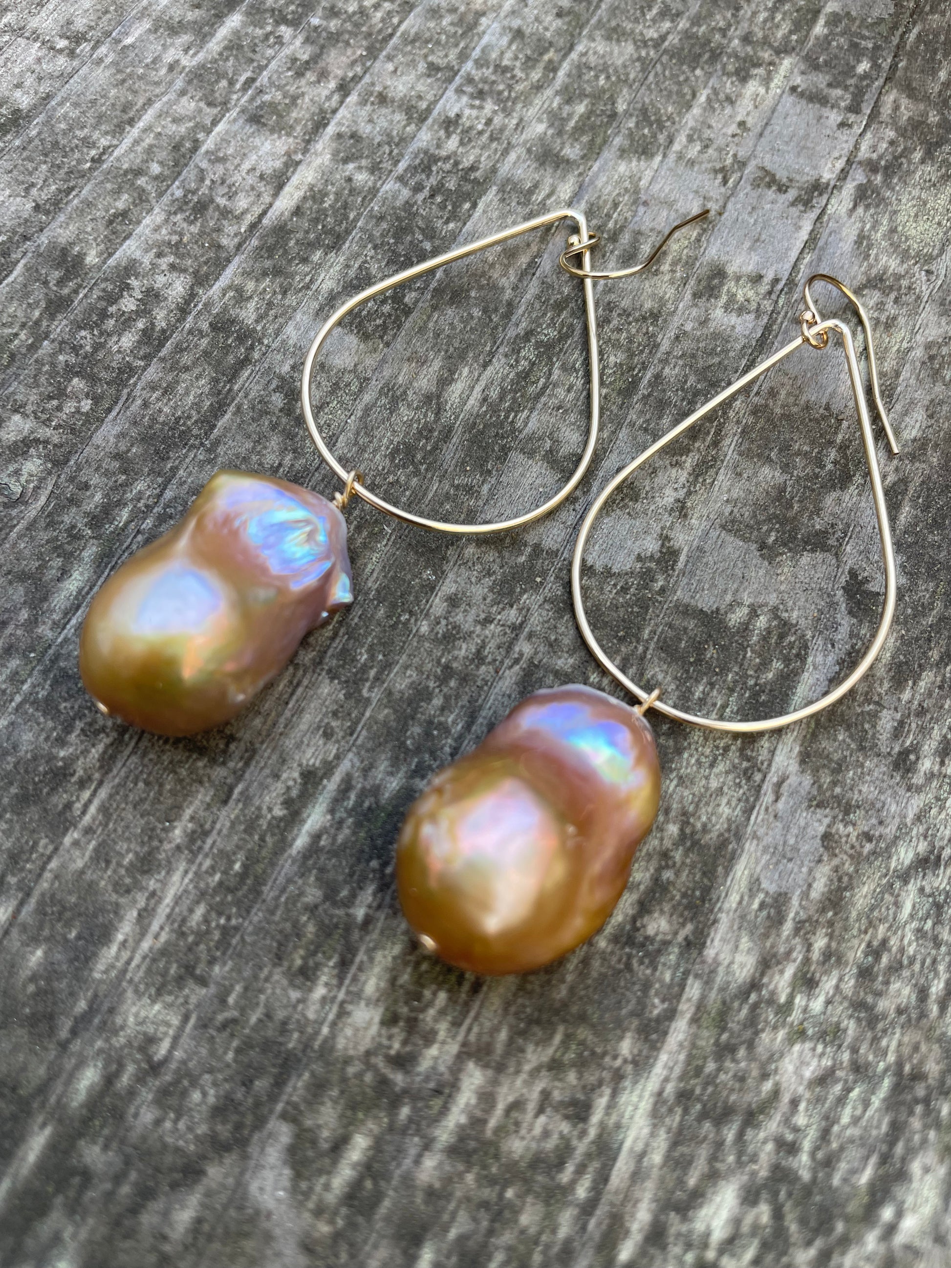 Large bronze baroque fireball pearls hanging off of a teardrop shaped gold wire with ear wires on a wooden background.