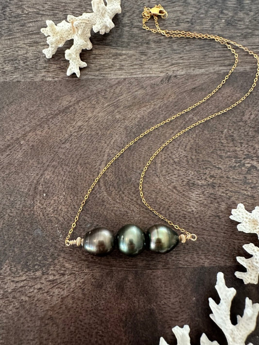 three black pearls in a row on a gold chain on a wooden background with white coral in the corners of the image