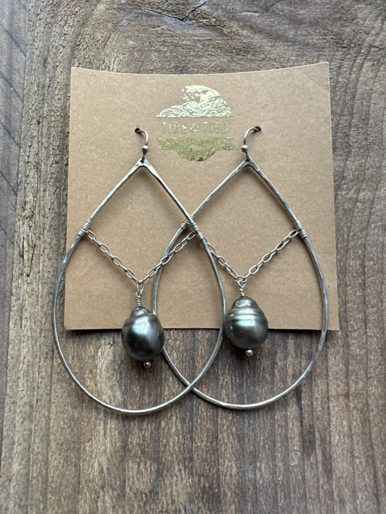 a pair of sterling silver earrings in a brown craft earring card wiht a gold logo with a chain suspending a black pearl. they are on a brown wooden background.