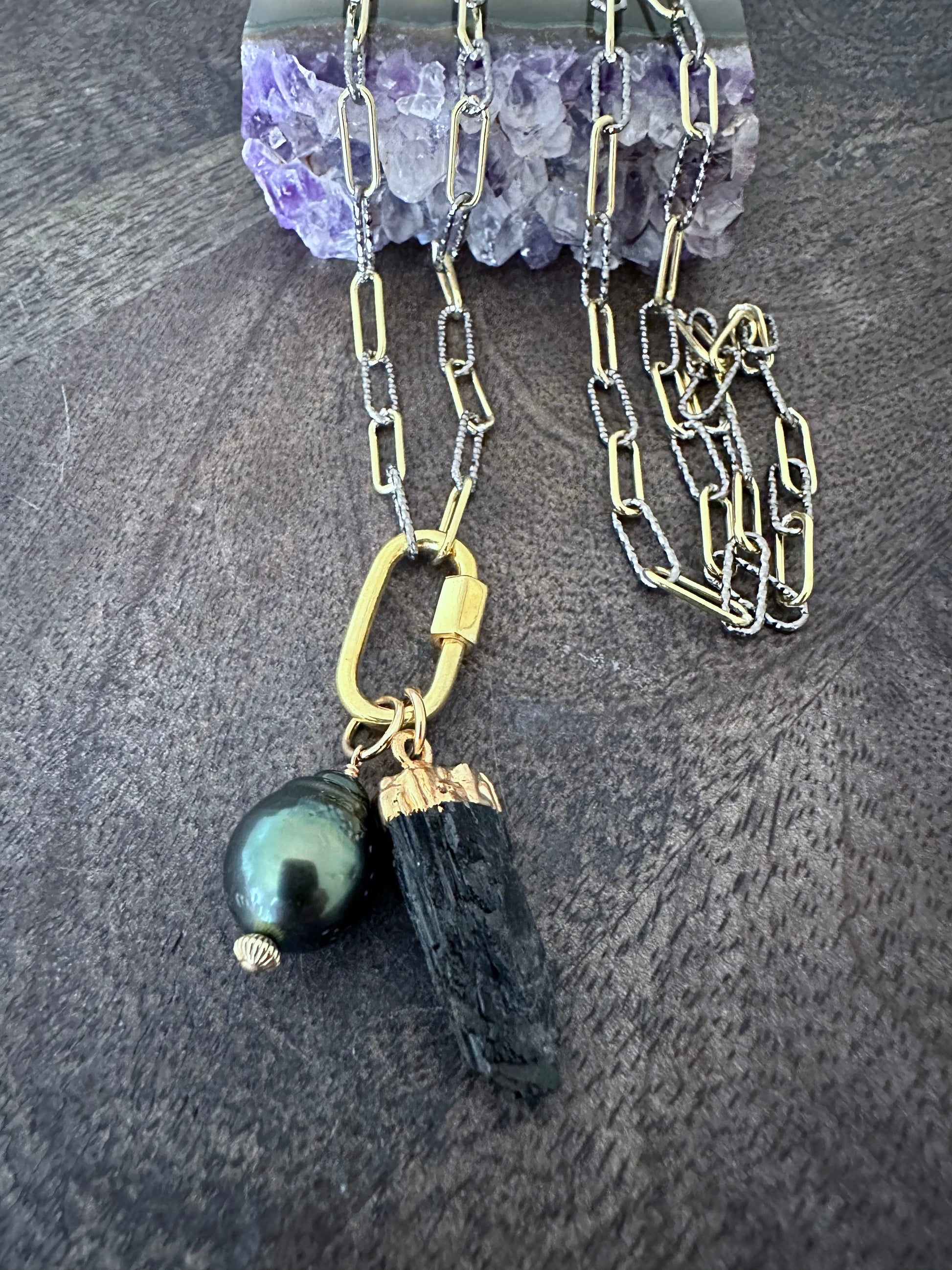 a long necklace wth mixed metal chain with a gold carabeaner and a large black pearl and a raw black tourmaline pendant on a purple amethyst chunk on a wooden background
