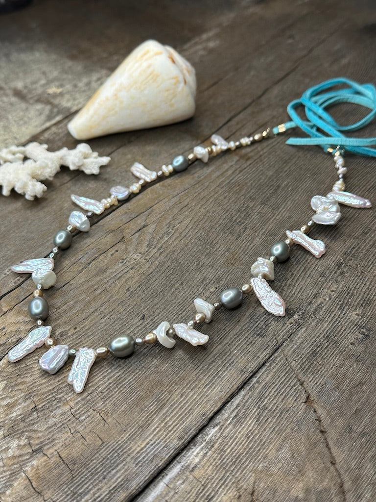 a close up of a necklace with turquoise suede tails and grey and silver pearls sits on a wooden background. a large shell is in the upper left corner