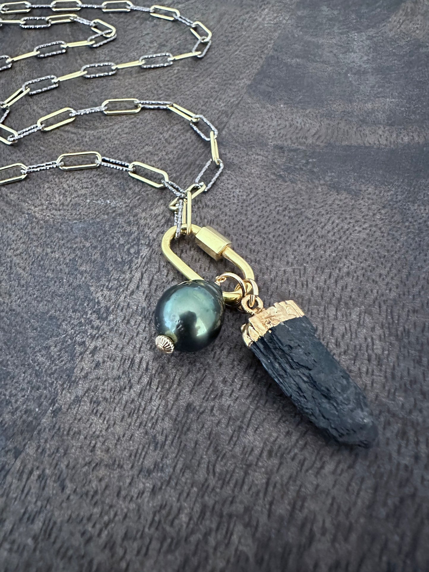 a close up of a long necklace wth mixed metal chain with a gold carabeaner and a large black pearl and a raw black tourmaline pendant on a wooden background