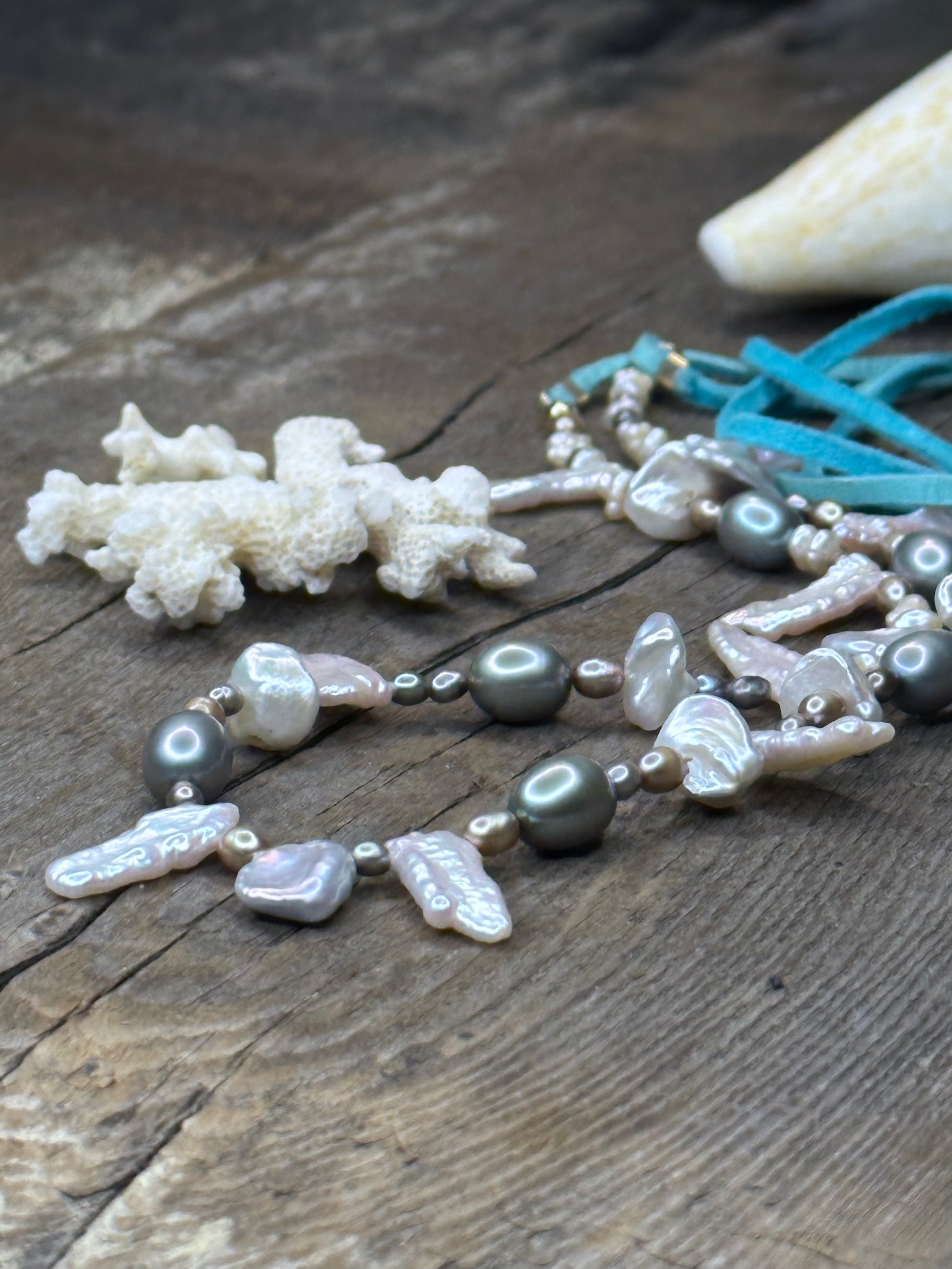 a close up of a close up of a necklace with turquoise suede tails and grey and silver pearls sits on a wooden background. a large shell is in the upper rt corner and a piece of white coral in the middle of the necklace