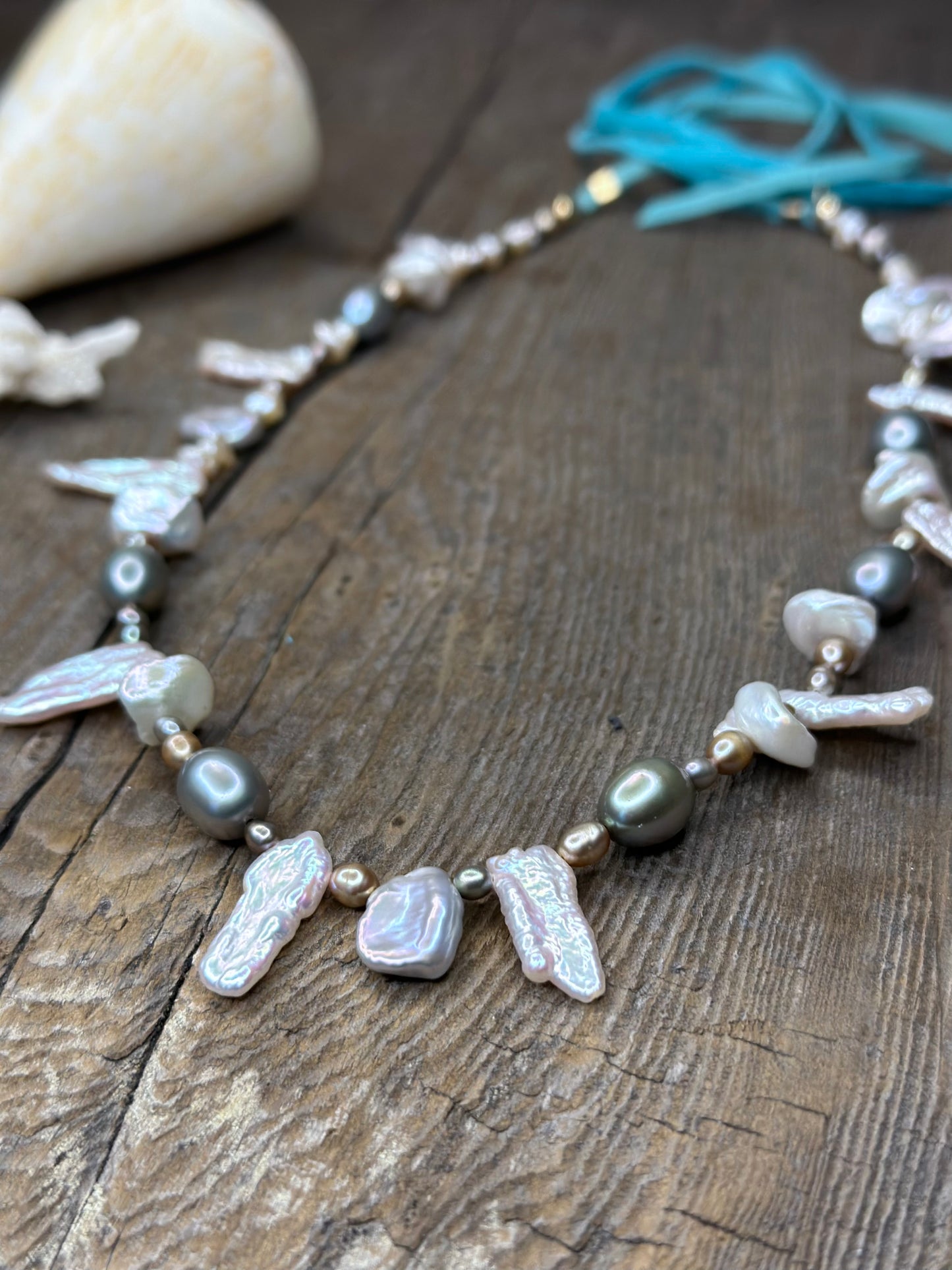 a close up of a necklace with turquoise suede tails and grey and silver pearls sits on a wooden background. a large shell is in the upper left corner