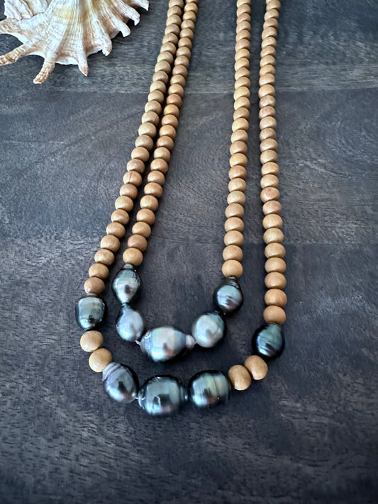 two necklaces layered on a wooden background. they both have wooden beads and several balck pearls. there is a shell in the upper left corner of the image.